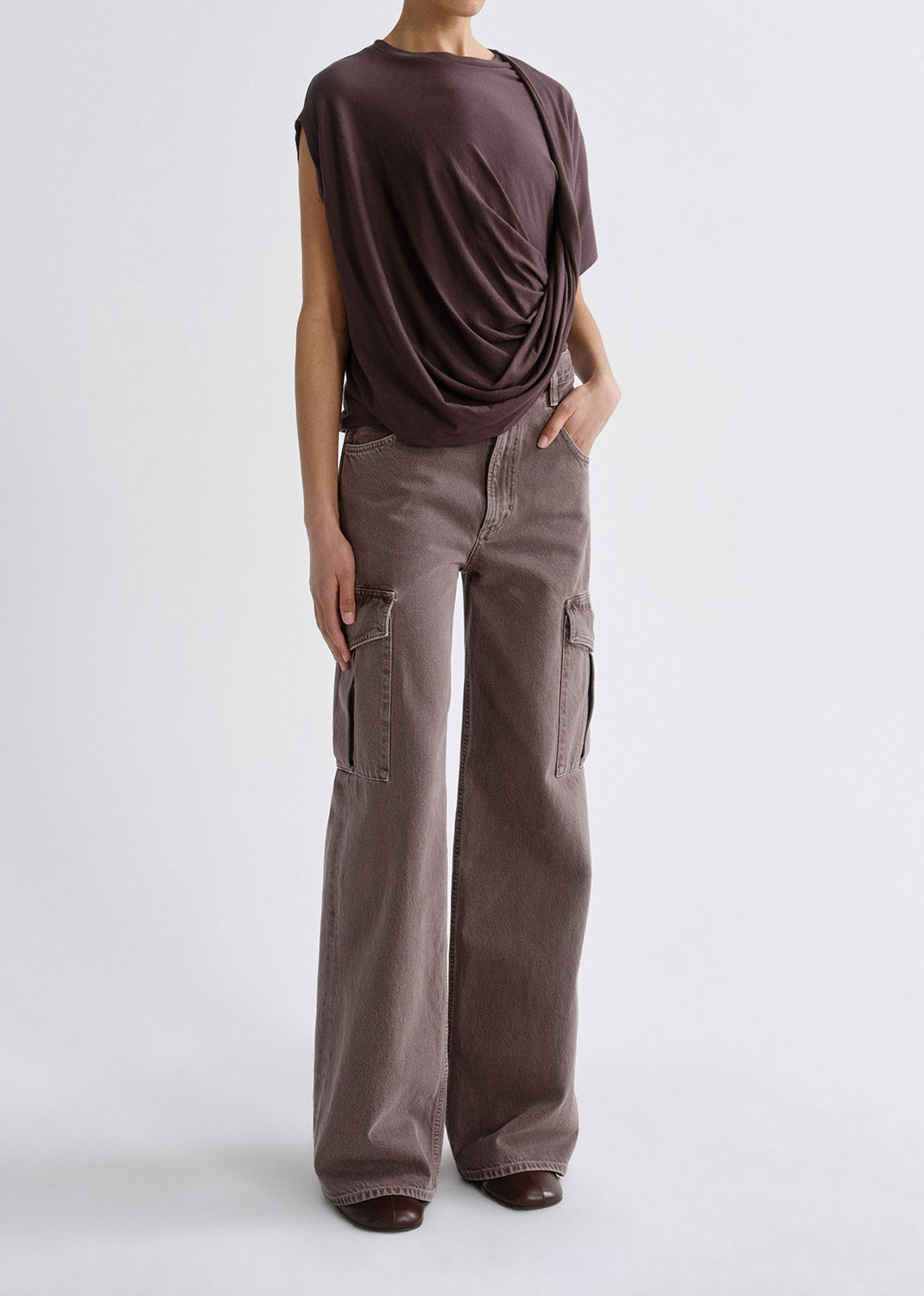 A model wearing a dark brown draped sleeveless top and wide-leg taupe cargo pants, standing against a grey background.