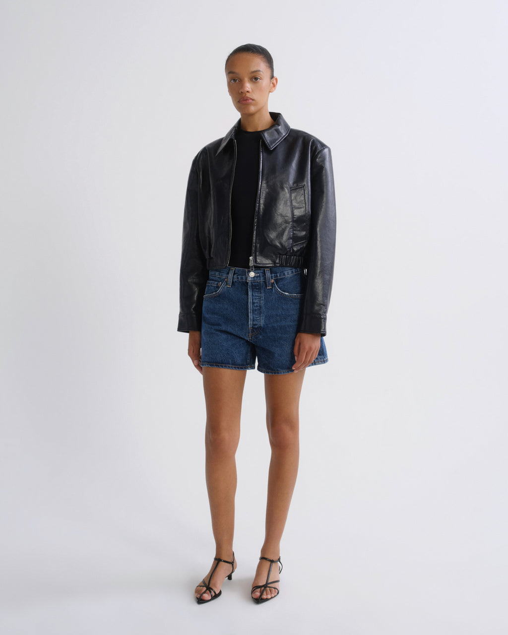 A model stands against a light gray studio background wearing a black leather jacket, a black top, blue denim shorts, and black strappy heels.