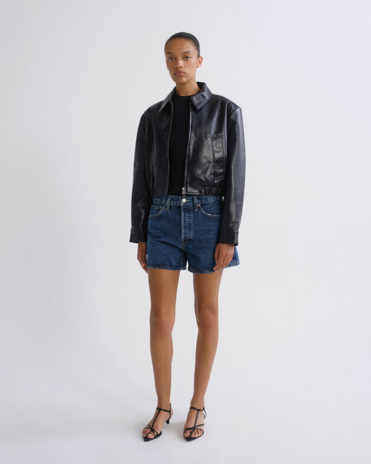 A model stands against a light gray studio background wearing a black leather jacket, a black top, blue denim shorts, and black strappy heels.
