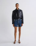 A model stands against a light gray studio background wearing a shiny black cropped leather jacket, blue denim shorts, and black strappy heels.
