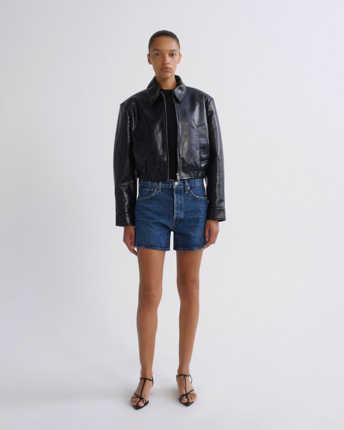 A model stands against a light gray studio background wearing a shiny black cropped leather jacket, blue denim shorts, and black strappy heels.