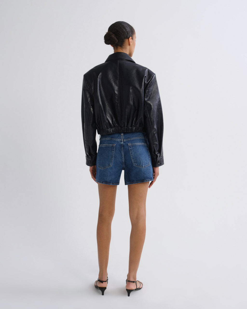 A model viewed from the back wearing a black faux-leather cropped jacket and blue denim shorts, standing against a plain light gray studio background.