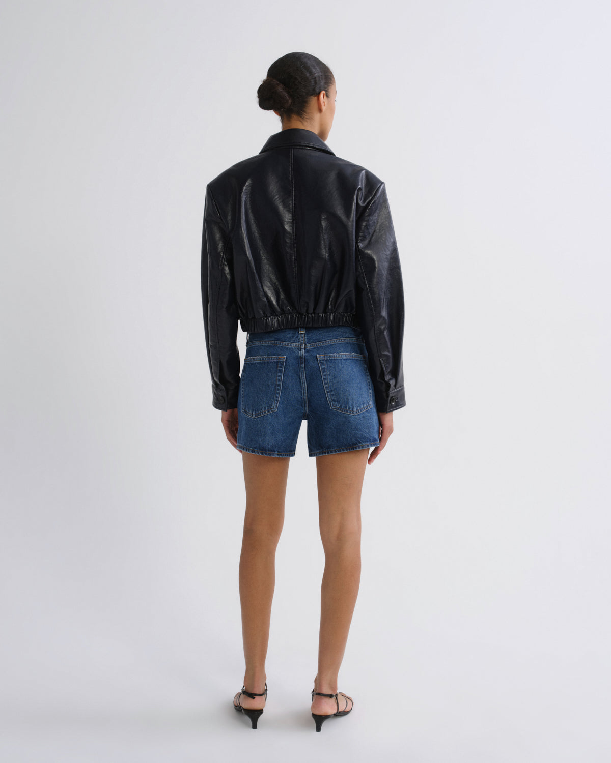 A model viewed from the back wearing a black faux-leather cropped jacket and blue denim shorts, standing against a plain light gray studio background.