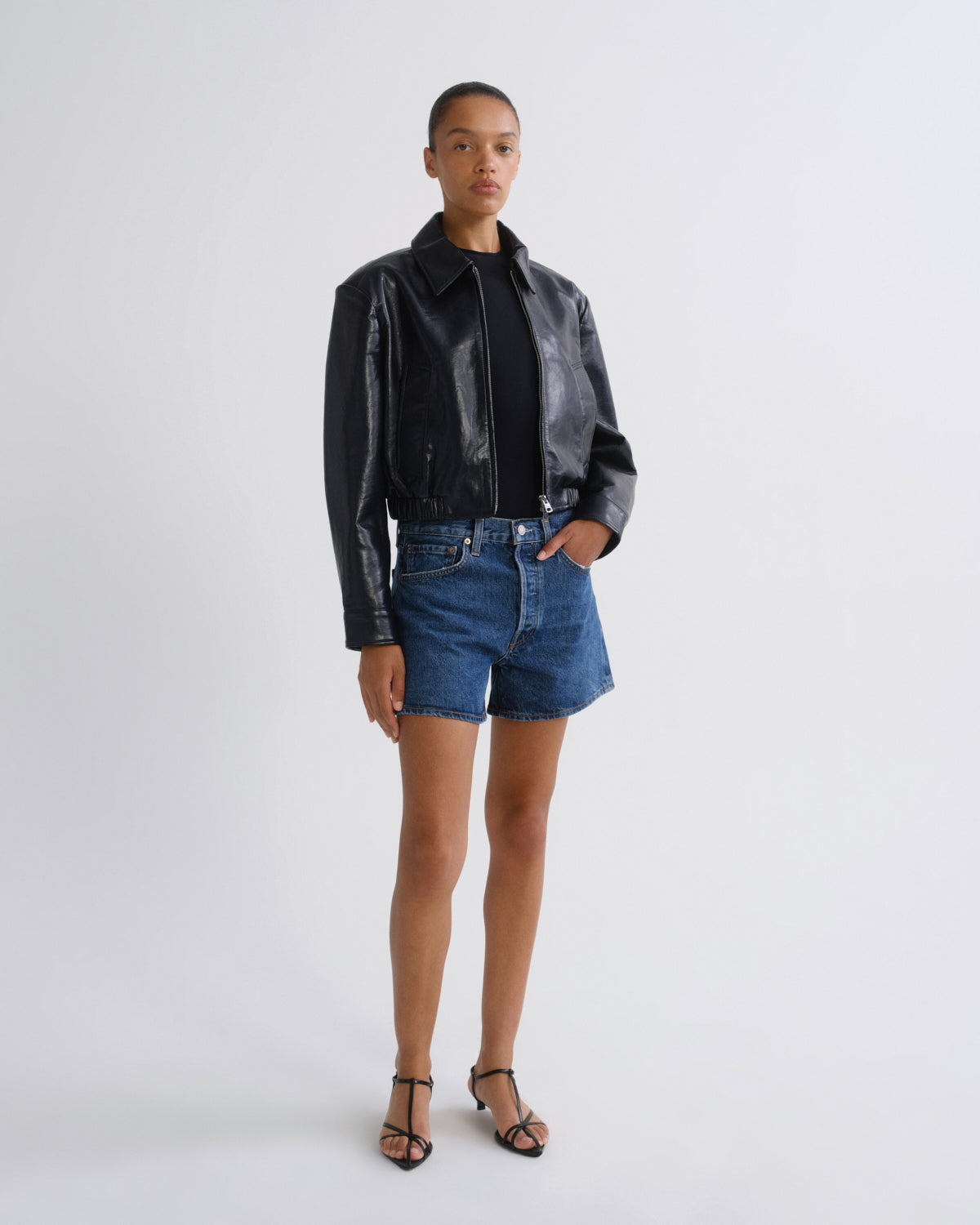 A model stands against a light gray studio background wearing a shiny black leather jacket over a black top, blue denim shorts, and black strappy heels.