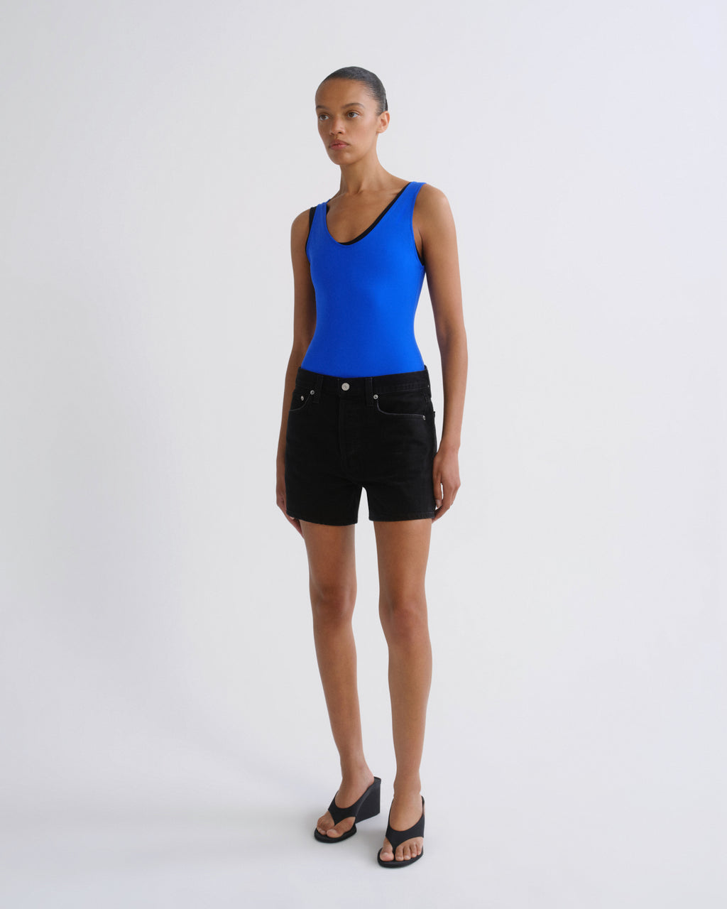 A model stands against a light gray studio background wearing a bright blue sleeveless top with black trim, black denim shorts, and black wedge sandals.