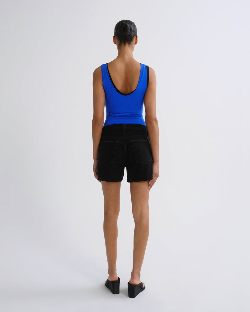 A model viewed from the back wearing a bright blue scoop-neck tank top and black shorts, standing against a plain light gray studio background.