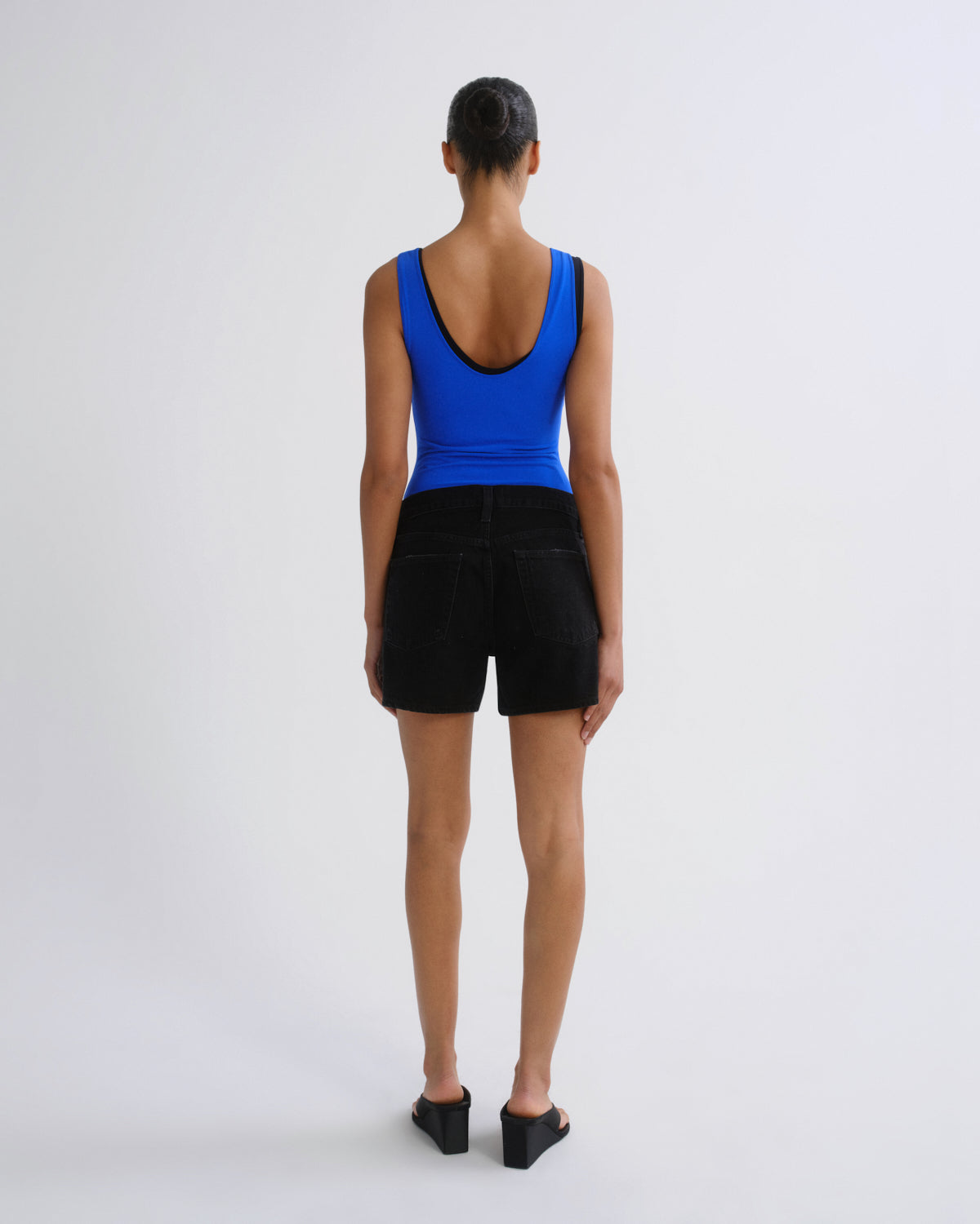 A model viewed from the back wearing a bright blue scoop-neck tank top and black shorts, standing against a plain light gray studio background.
