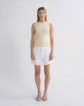 A model stands against a light gray studio background wearing a pale yellow ribbed sleeveless top, white denim shorts, and clear-strap high-heeled sandals.