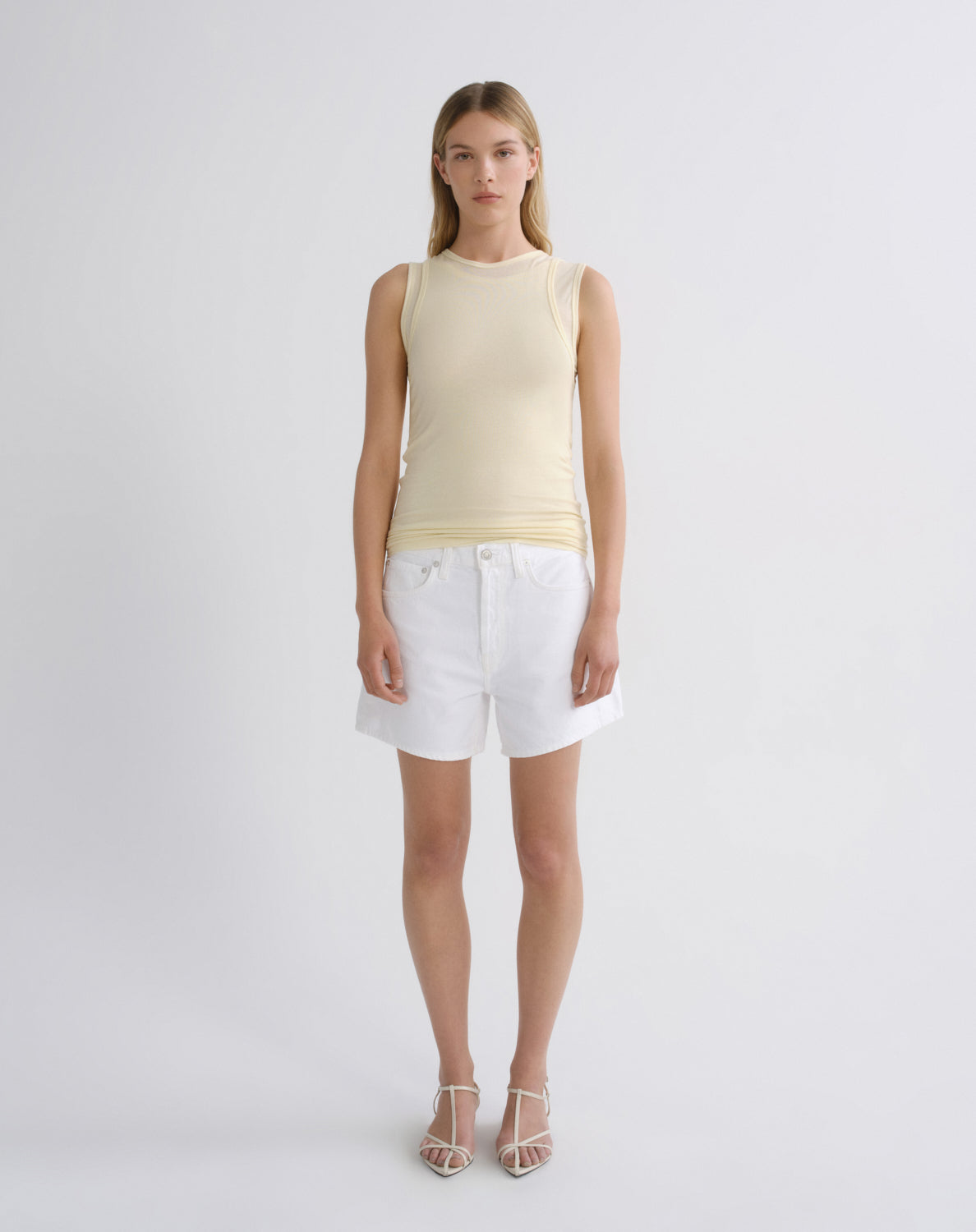 A model stands against a light gray studio background wearing a pale yellow ribbed sleeveless top, white denim shorts, and clear-strap high-heeled sandals.