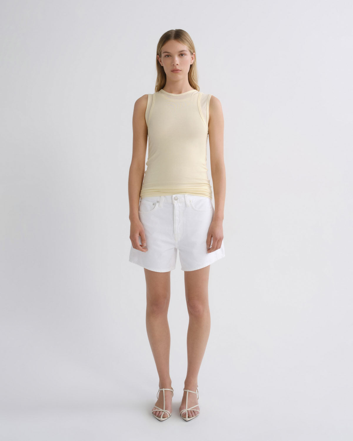 A model stands against a light gray studio background wearing a pale cream ribbed sleeveless top, white denim shorts, and clear-strap high-heeled sandals.