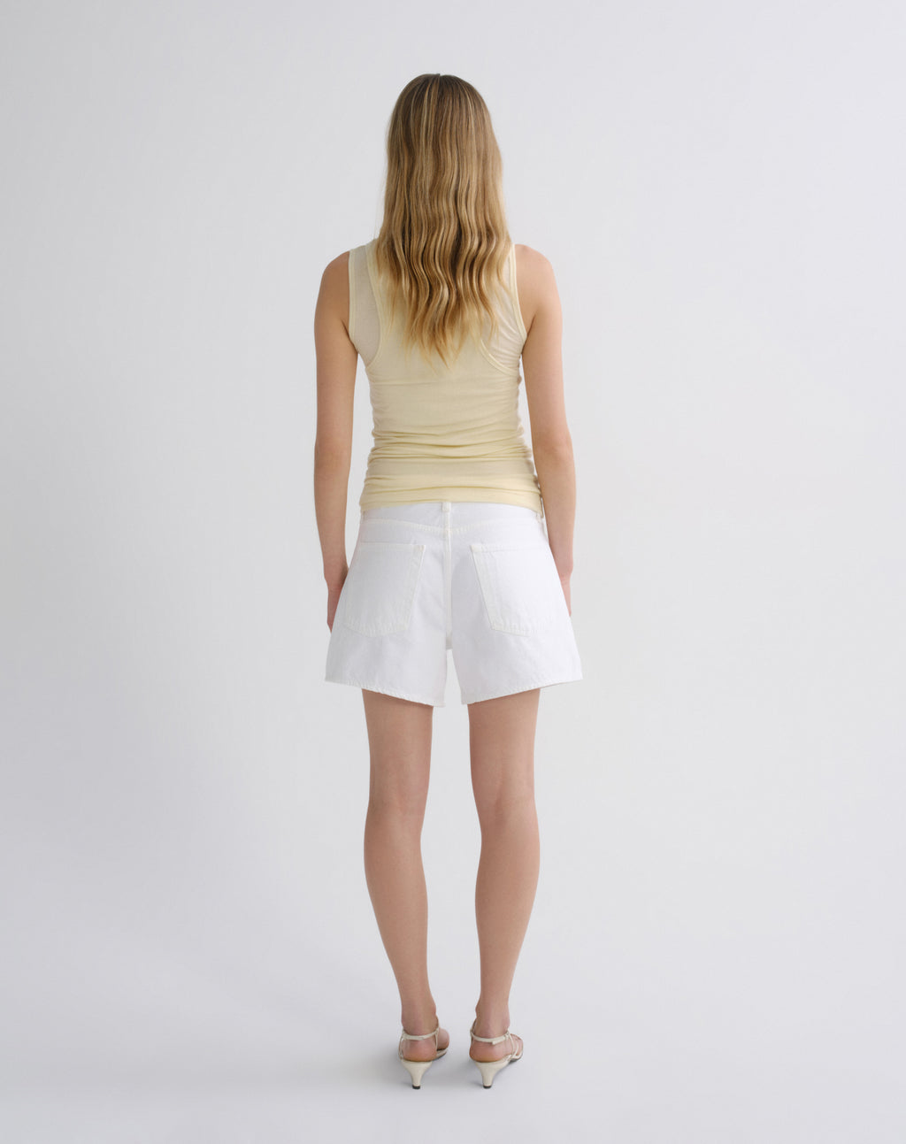A woman viewed from behind wearing a pale yellow sleeveless top and white shorts, standing against a plain light gray studio background.