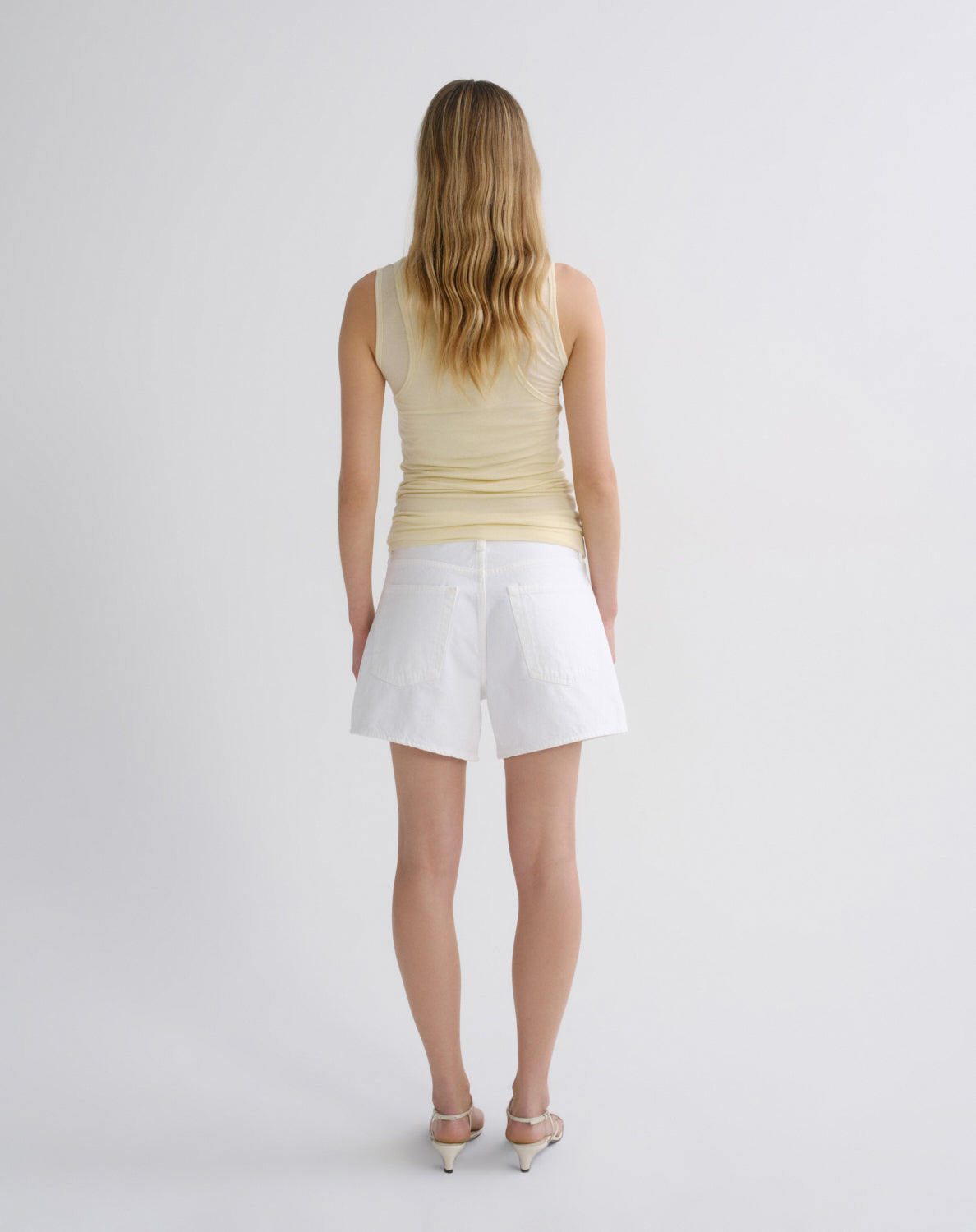 A woman viewed from behind wearing a pale yellow sleeveless top and white shorts, standing against a plain light gray studio background.