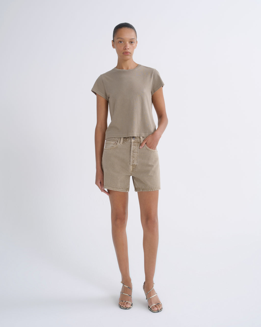 A model stands against a light gray studio background wearing a taupe short-sleeve crewneck t-shirt, matching beige denim shorts, and beige strappy high-heeled sandals.