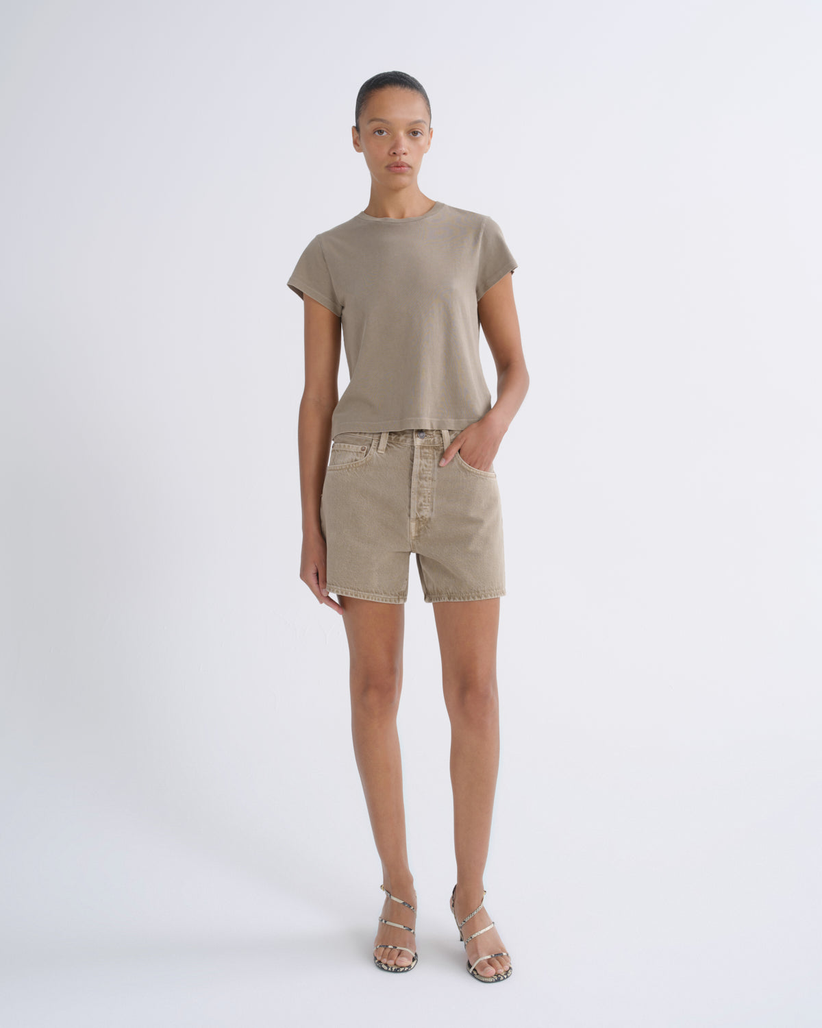 A model stands against a light gray studio background wearing a taupe short-sleeve crewneck t-shirt, matching beige denim shorts, and beige strappy high-heeled sandals.
