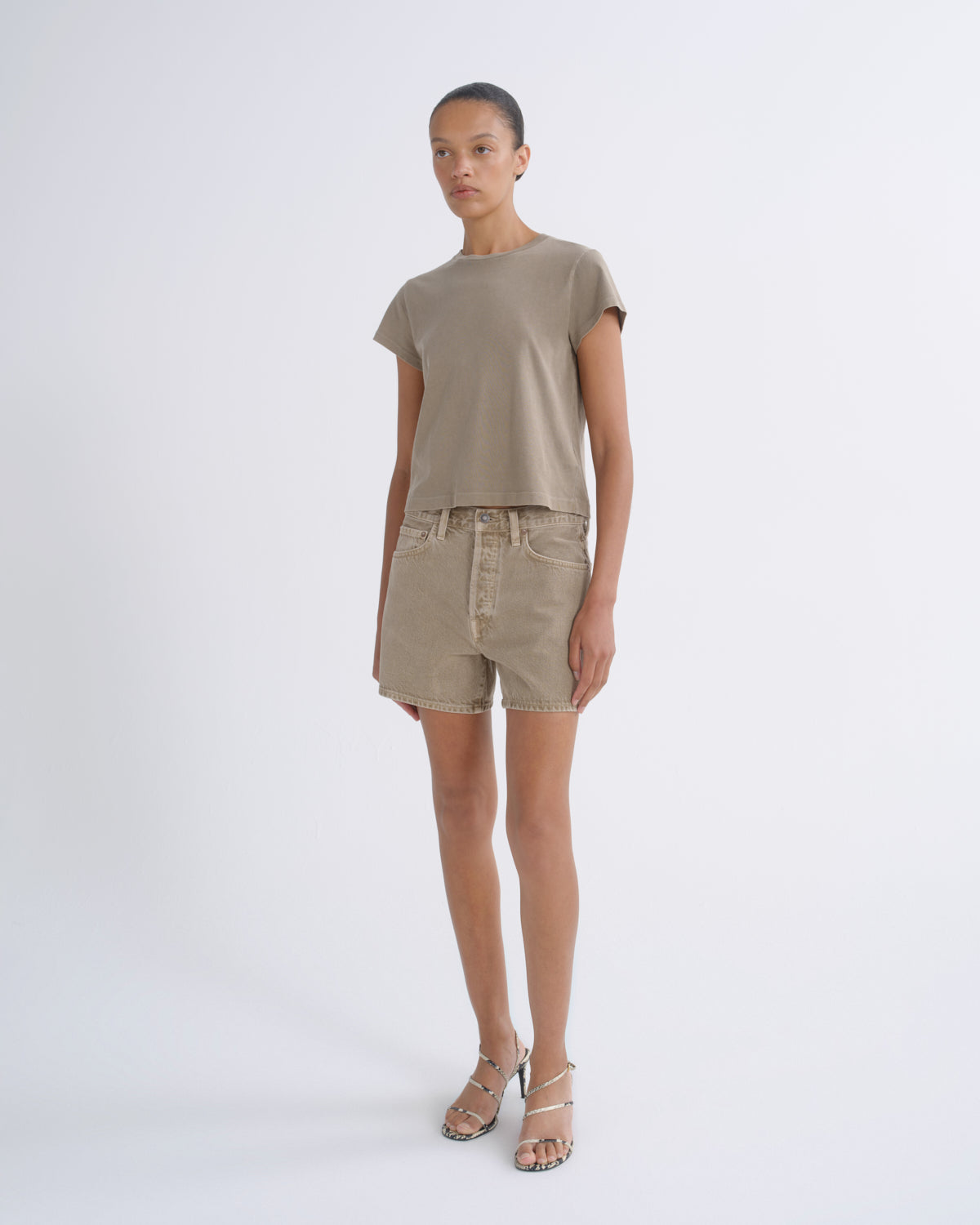 A model stands against a light gray studio background wearing a taupe short-sleeve cropped T-shirt and matching beige shorts, paired with metallic heeled sandals.