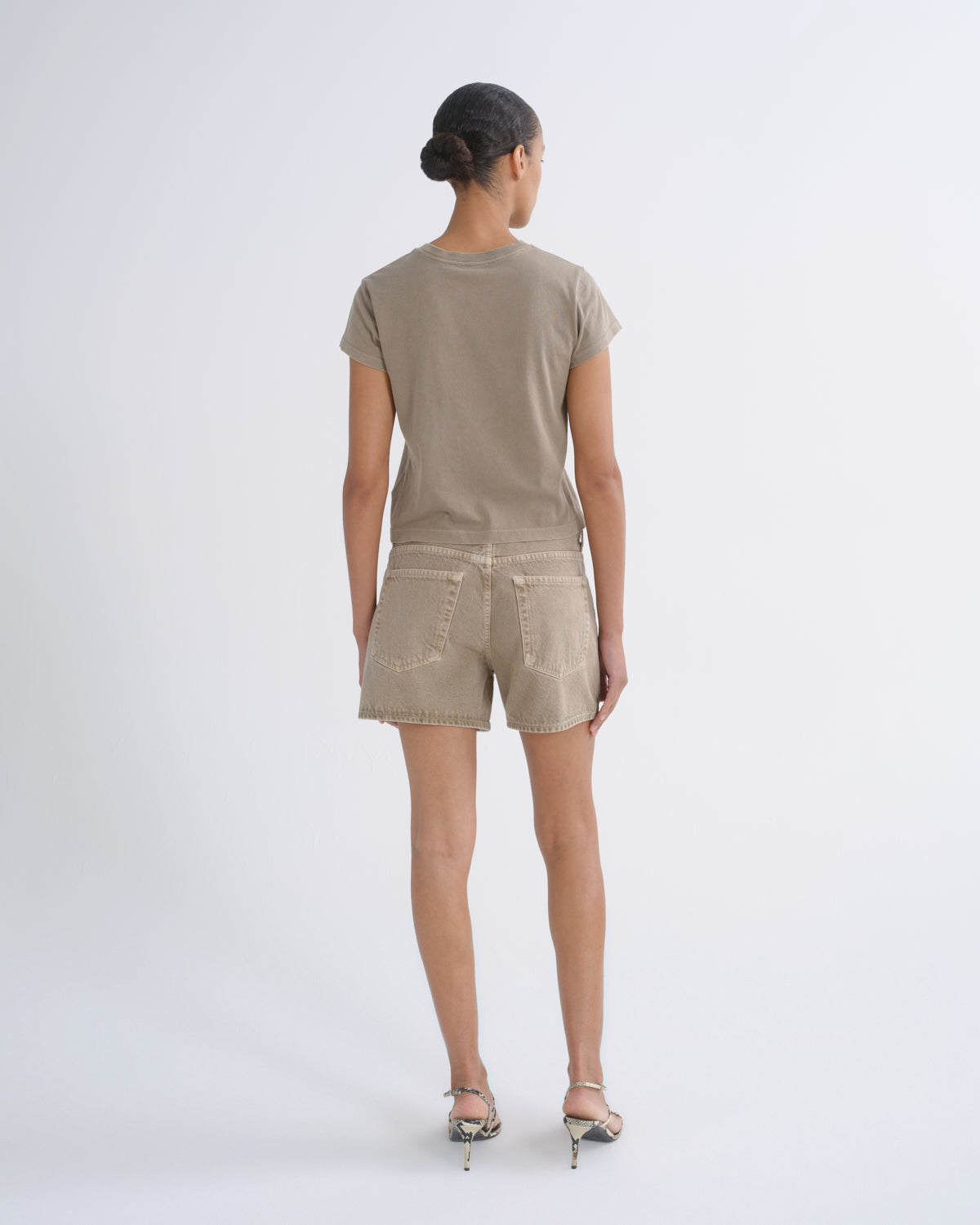 A model viewed from behind wearing a taupe short-sleeve t-shirt and matching beige denim shorts, paired with metallic high-heeled sandals, against a plain light gray studio background.