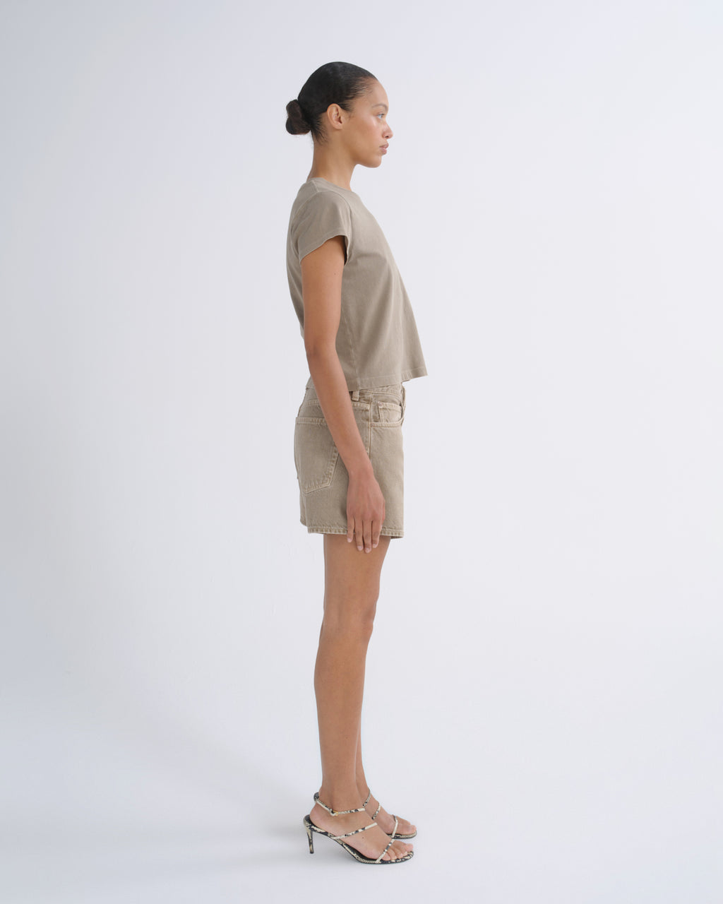 A model in side profile wearing a matching beige short-sleeve crop top and shorts set with metallic heeled sandals, photographed against a seamless light gray studio background.
