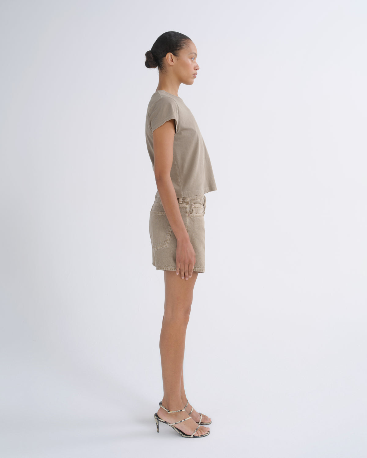 A model in side profile wearing a matching beige short-sleeve crop top and shorts set with metallic heeled sandals, photographed against a seamless light gray studio background.