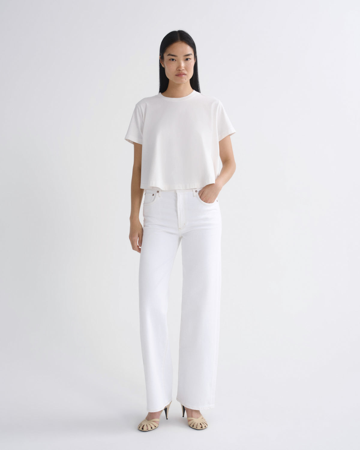 A model stands against a light gray studio background wearing a white short-sleeve cropped T-shirt, white wide-leg pants, and beige heeled sandals.