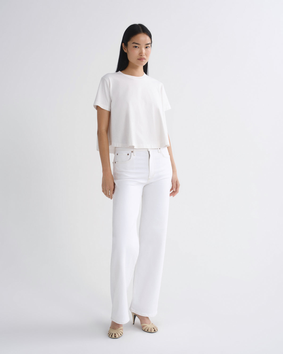 A model stands against a light gray studio background wearing a white short-sleeve cropped top, white wide-leg pants, and beige heeled sandals.