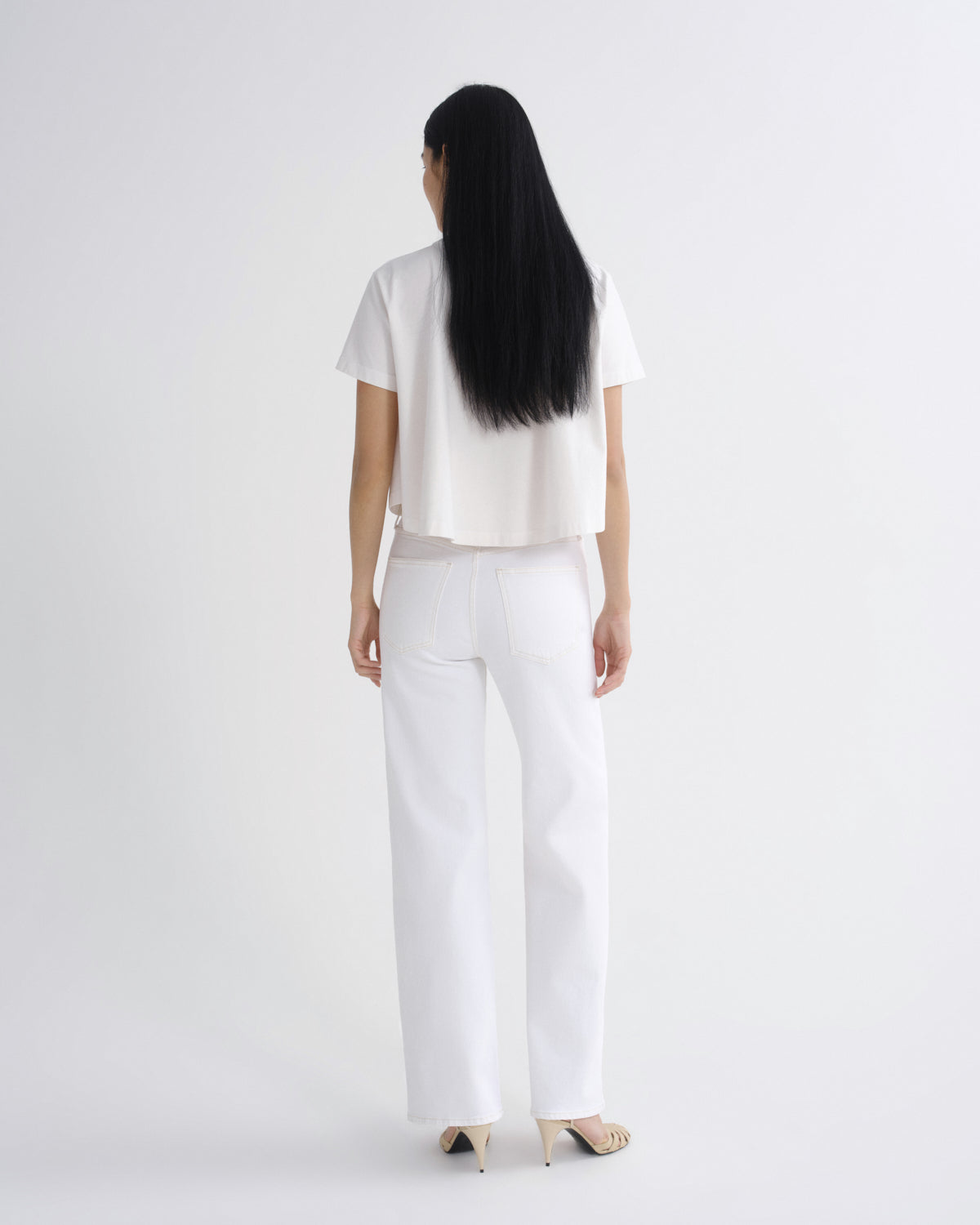 A woman viewed from behind wearing a white short-sleeve cropped top and white wide-leg trousers, paired with beige high-heeled sandals, against a plain light gray studio background.