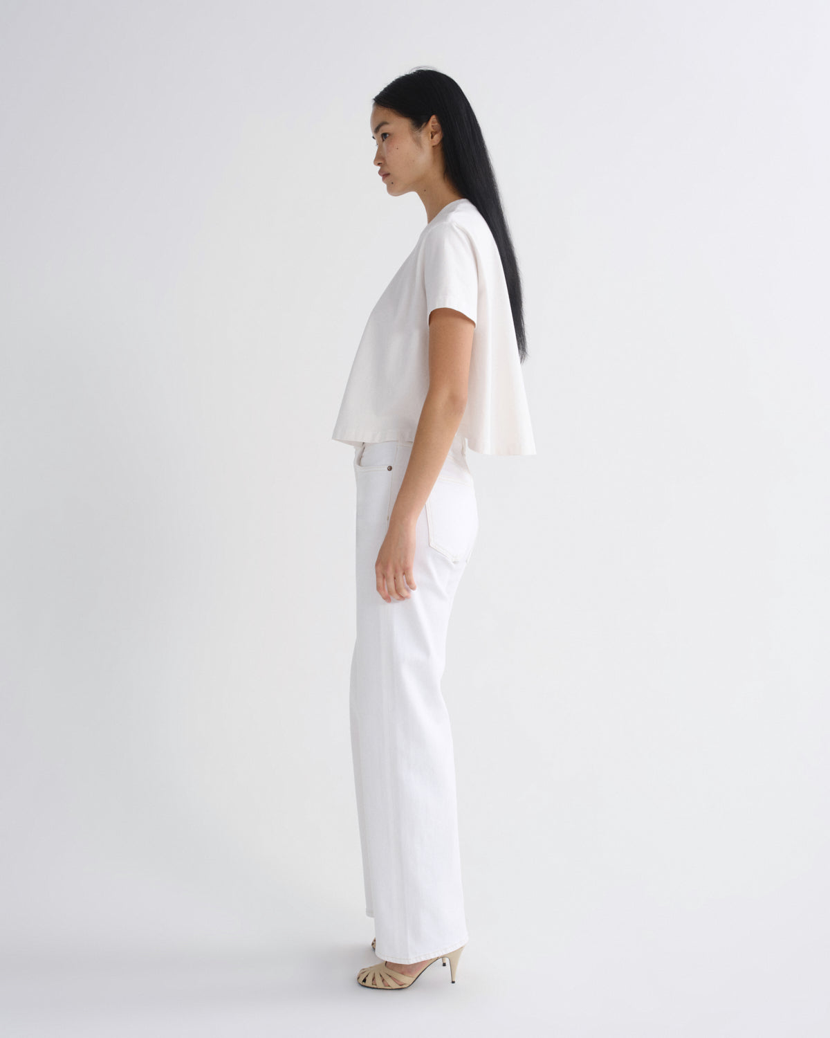 A woman in a side profile wearing a white short-sleeve top with a draped cape detail and matching white wide-leg trousers, paired with beige high-heeled sandals, against a plain white studio background.