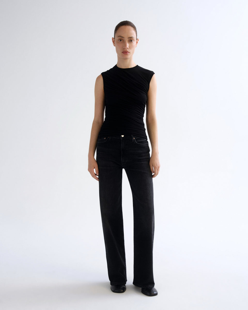 A model stands against a plain white studio background wearing a sleeveless black top with draped detailing and black wide-leg jeans.
