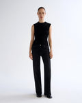 A model stands against a plain white studio background wearing a sleeveless black top with draped detailing and black wide-leg jeans.