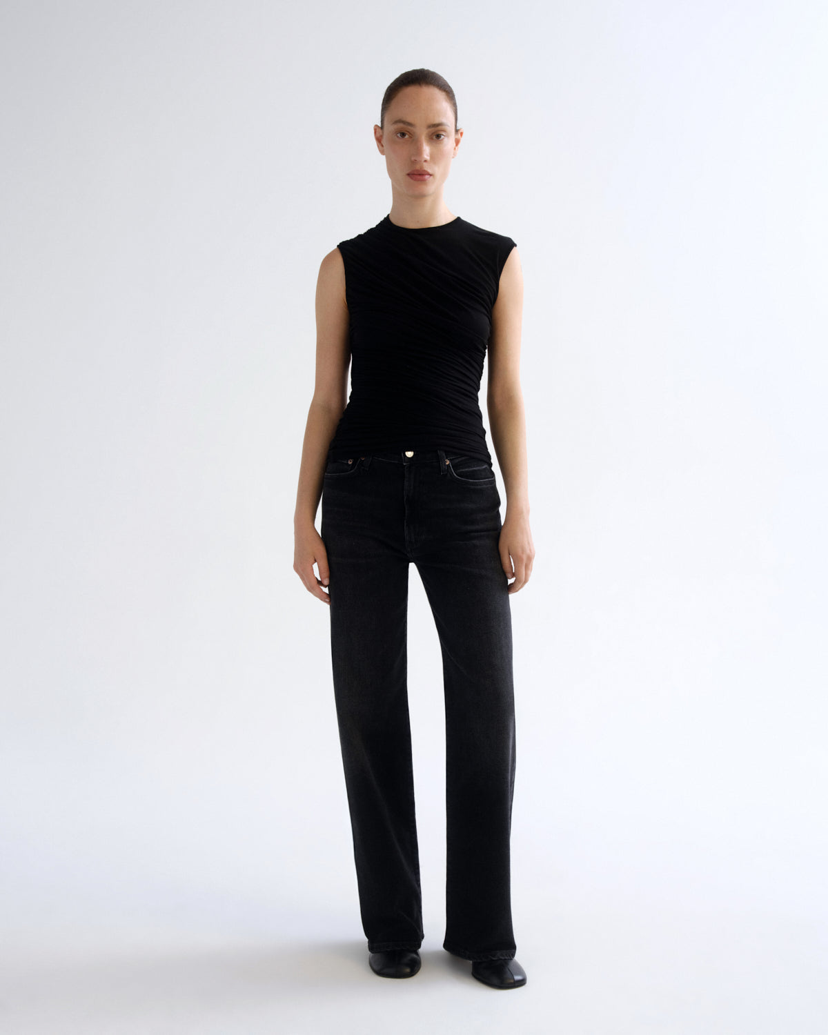 A model stands against a plain white studio background wearing a sleeveless black top with draped detailing and black wide-leg jeans.