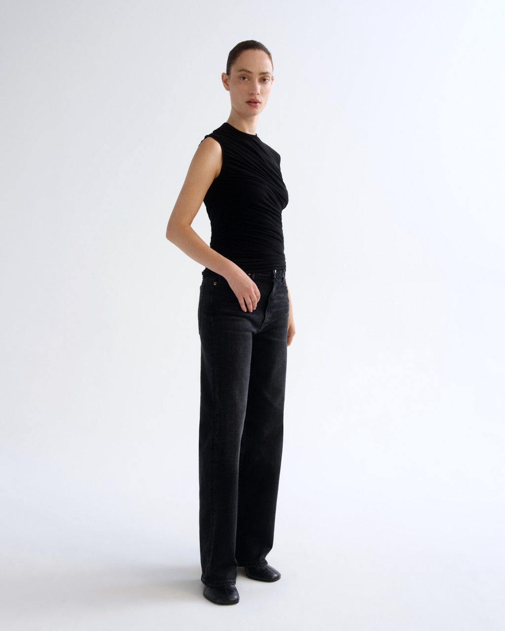 A model stands in profile wearing a sleeveless black ruched top and black wide-leg trousers against a clean white studio background.