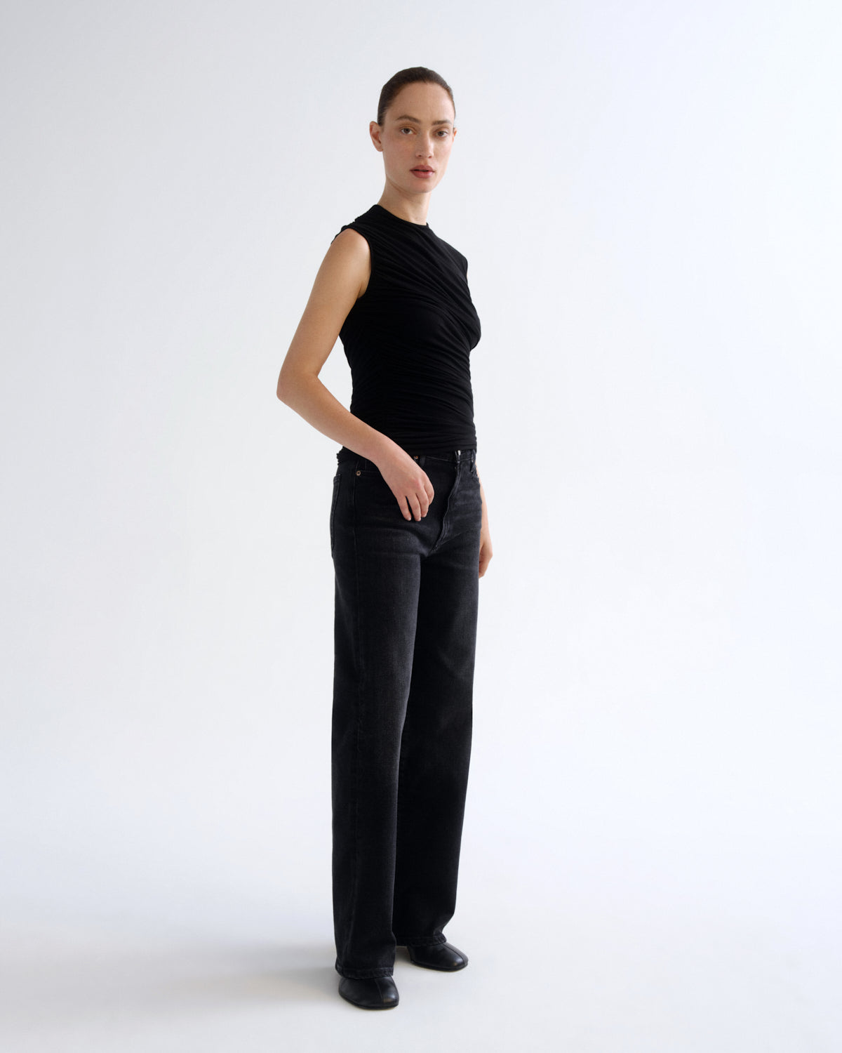 A model stands in profile wearing a sleeveless black ruched top and black wide-leg trousers against a clean white studio background.