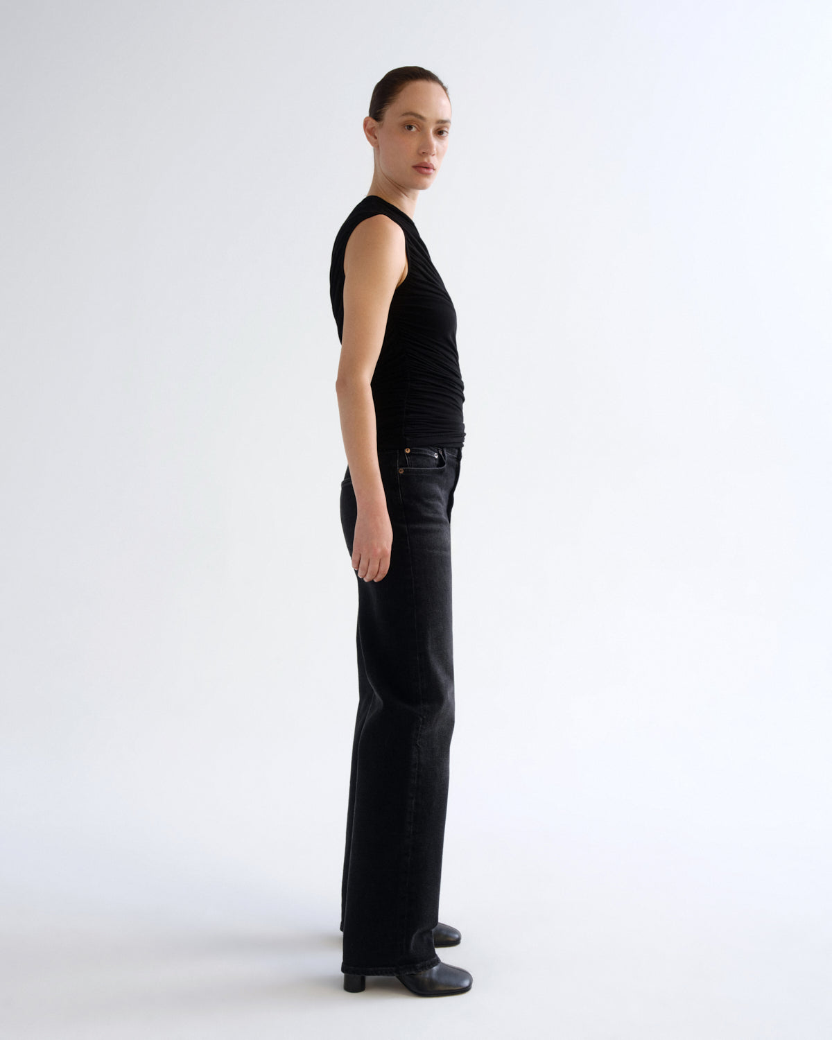 A model stands in profile wearing a black sleeveless top, black wide-leg jeans, and black shoes against a plain light gray studio background.