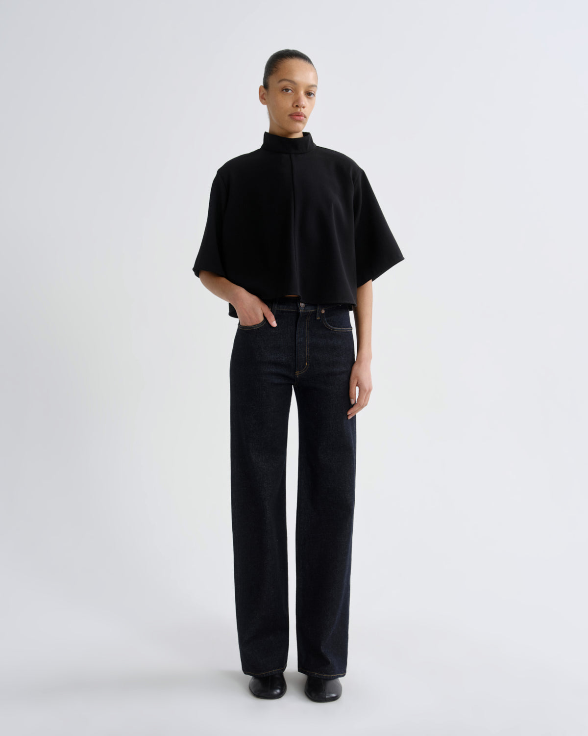 A model stands against a light gray studio background wearing a black short-sleeve mock-neck top and dark wide-leg jeans.