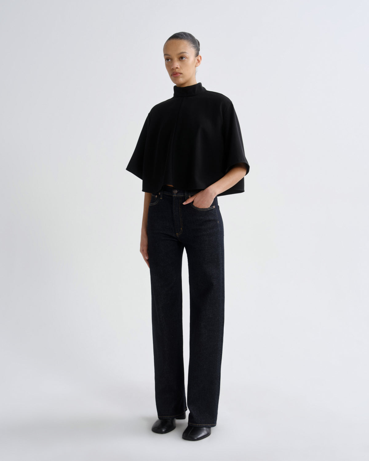 A model stands against a light gray studio background wearing a black short-sleeved mock-neck top, dark wide-leg jeans, and black shoes.