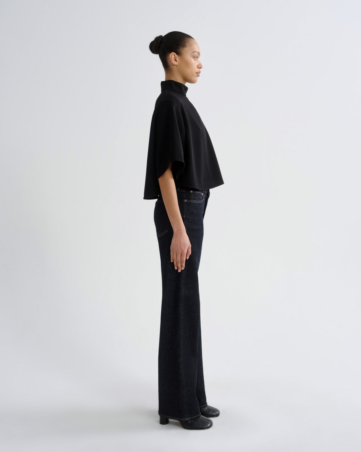 A model in profile wearing a black cropped cape-sleeve mock-neck top and dark wide-leg jeans, standing against a plain light gray studio background.