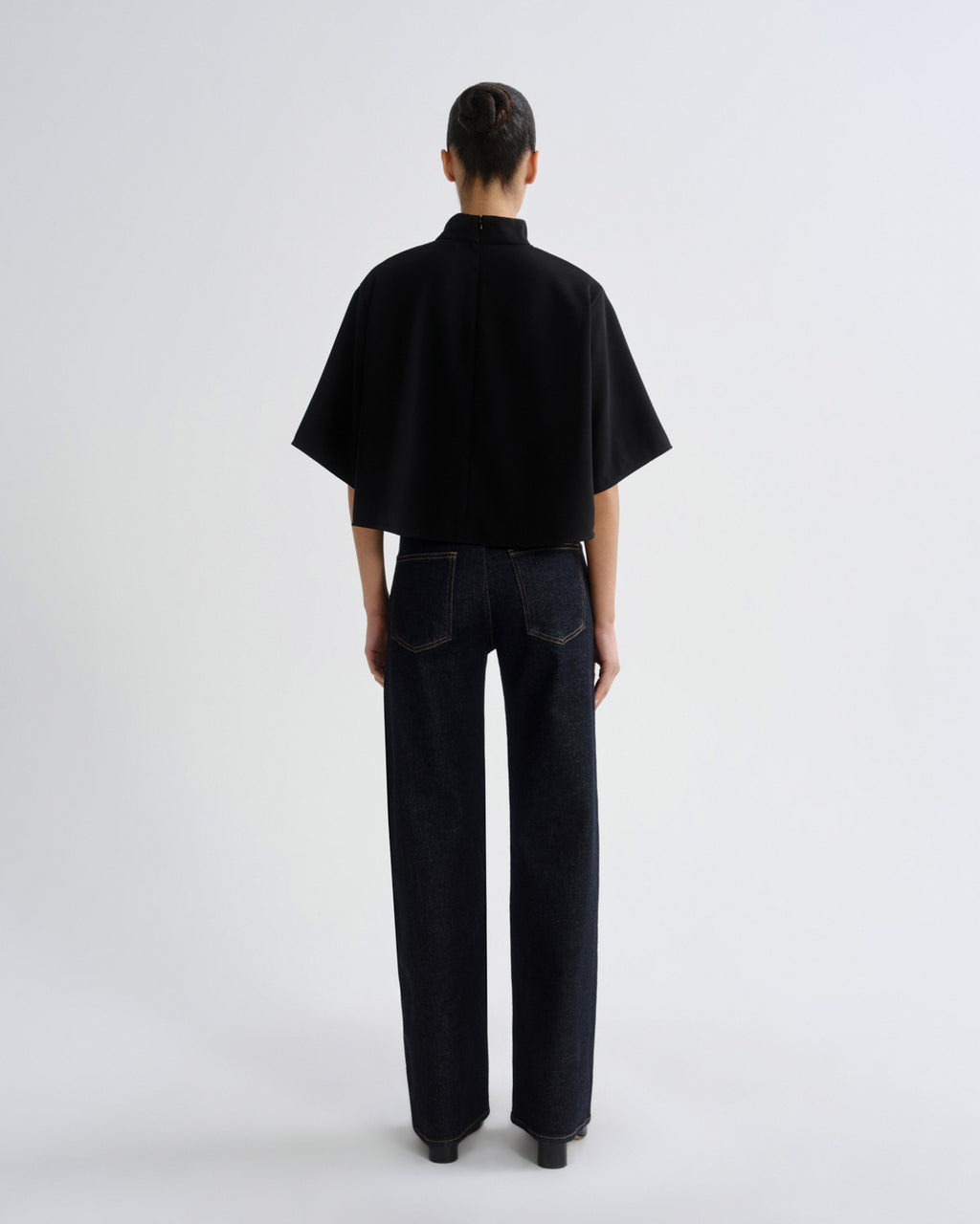 A model viewed from the back wearing a black short-sleeved high-neck top and dark wide-leg jeans against a plain light gray studio background.