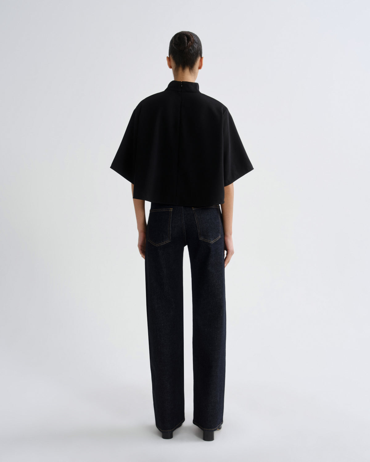 A model viewed from the back wearing a black short-sleeved high-neck top and dark wide-leg jeans against a plain light gray studio background.