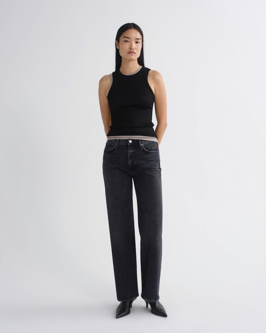 A model stands against a light gray studio background wearing a black sleeveless ribbed top, dark gray wide-leg jeans, and black pointed shoes.