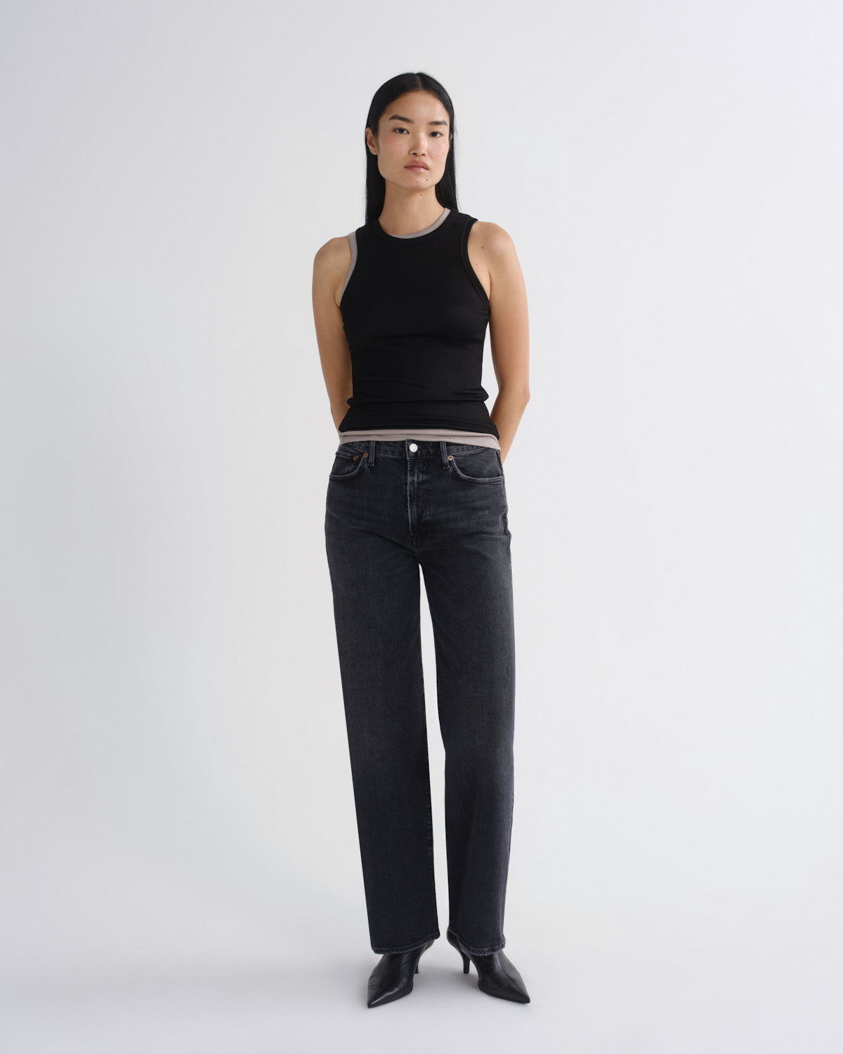 A model stands against a light gray studio background wearing a black sleeveless ribbed top, dark gray wide-leg jeans, and black pointed shoes.
