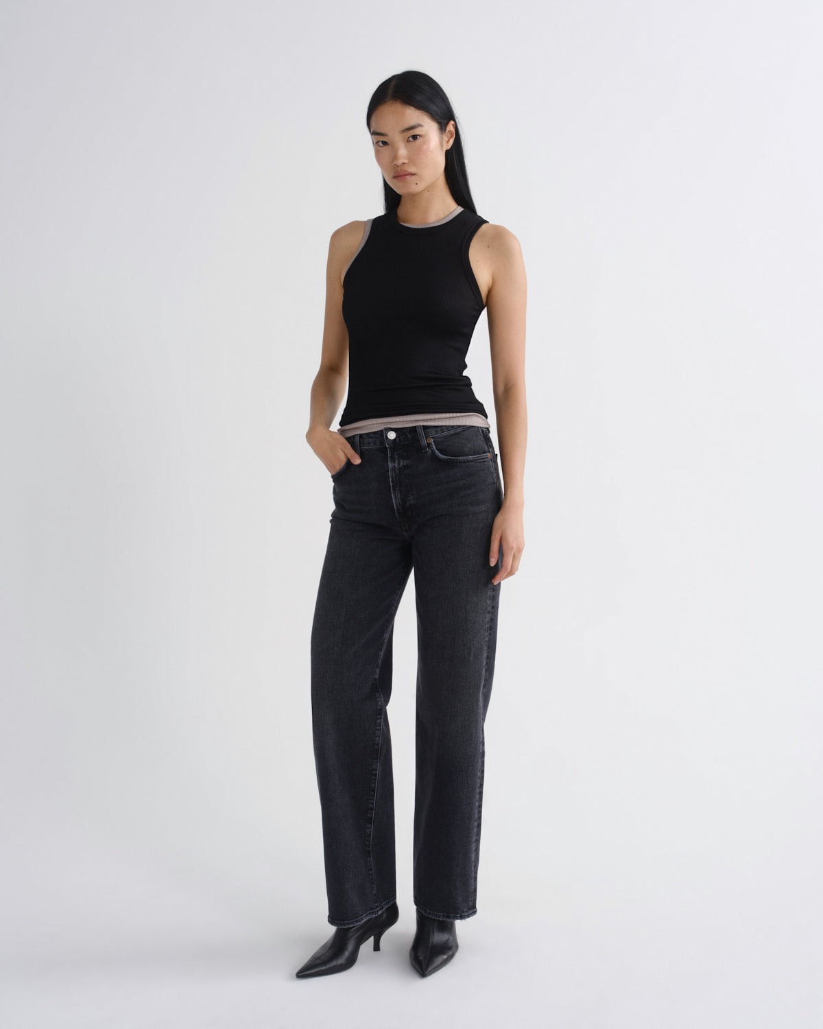 A model stands against a light gray studio background wearing a fitted black sleeveless top, dark charcoal straight-leg jeans, and black pointed-toe heels.