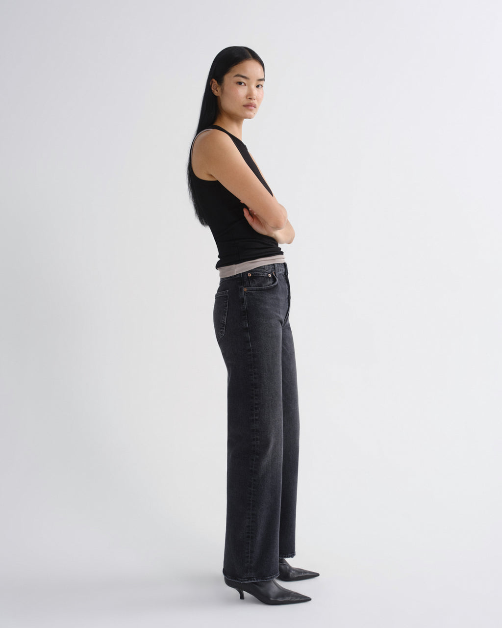 A woman stands in profile wearing a black sleeveless top, dark gray wide-leg jeans, and black pointed-toe heels against a plain light gray studio background.