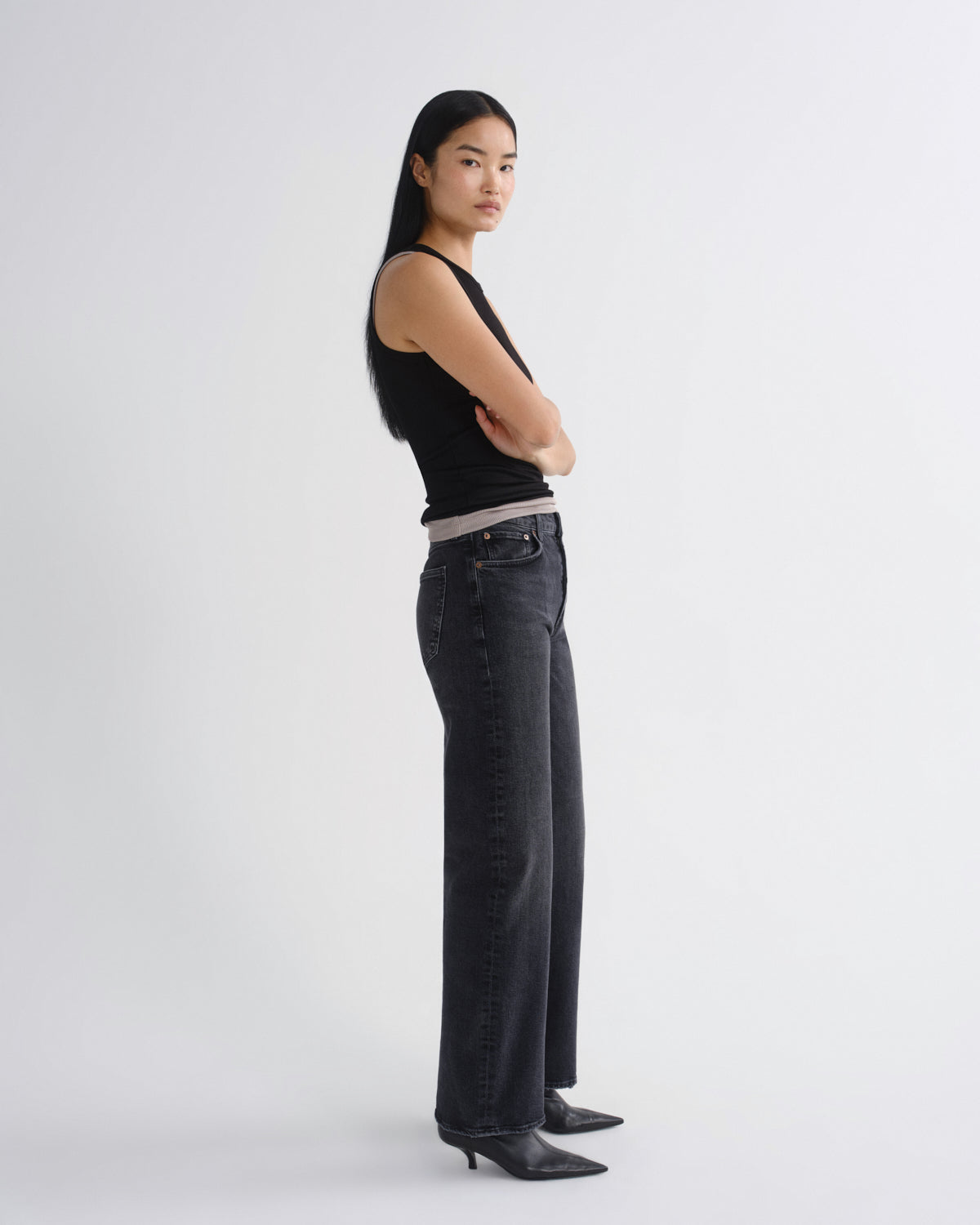 A woman stands in profile wearing a black sleeveless top, dark gray wide-leg jeans, and black pointed-toe heels against a plain light gray studio background.