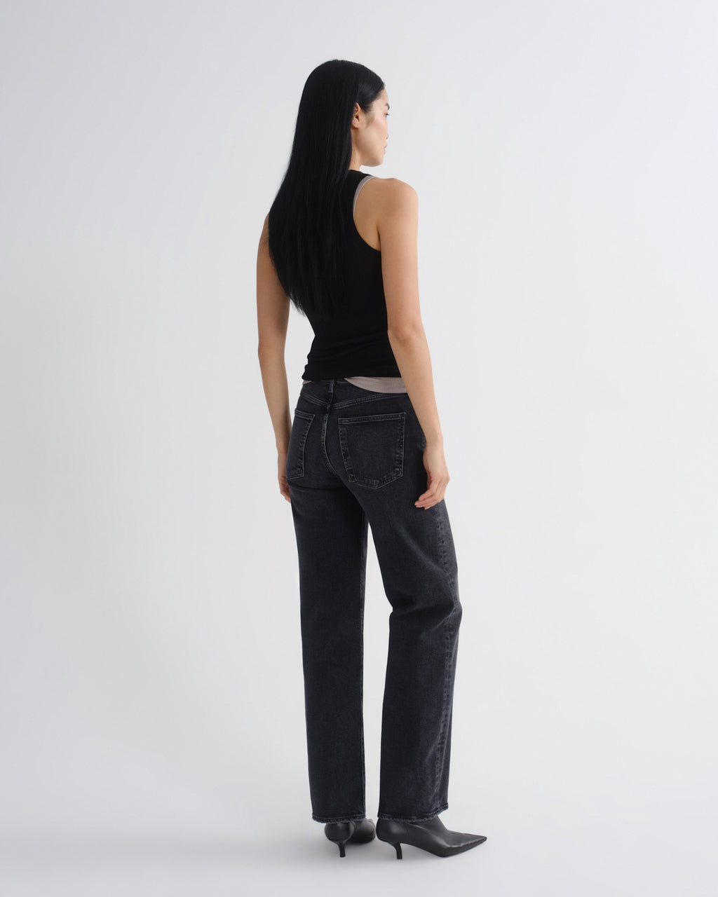 A model shown from the back wearing a black sleeveless top, dark gray wide-leg jeans, and black pointed-toe boots against a light gray studio background.