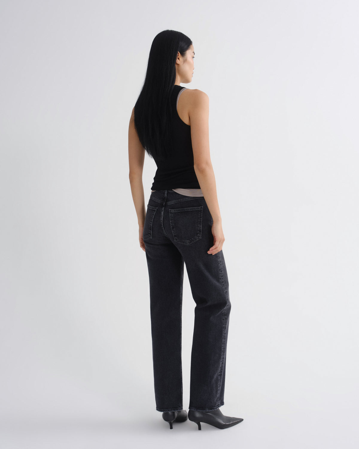 A model shown from the back wearing a black sleeveless top, dark gray wide-leg jeans, and black pointed-toe boots against a light gray studio background.