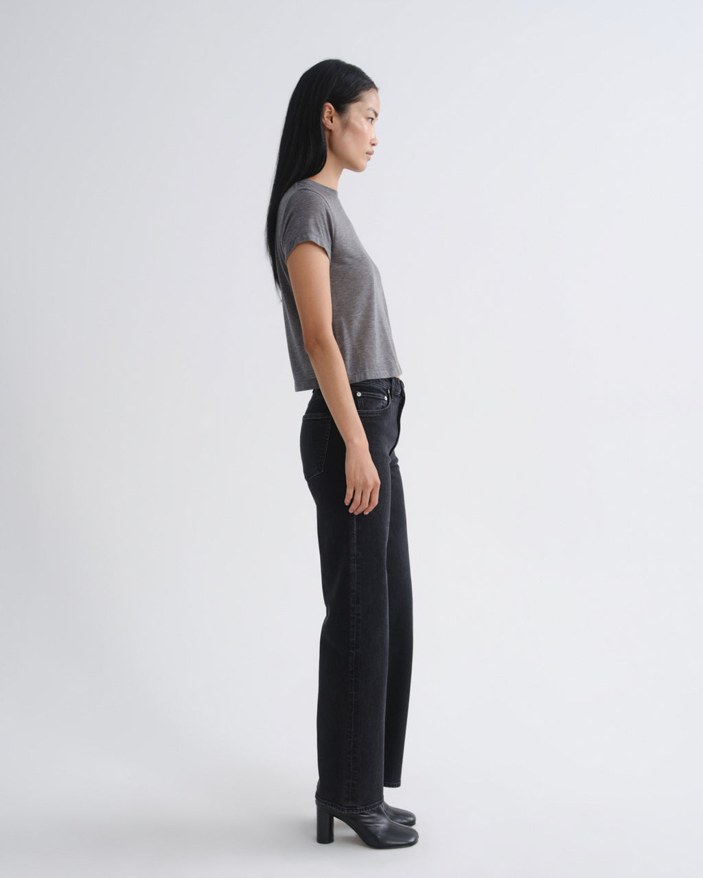 A woman stands in profile wearing a short-sleeve gray knit top, black wide-leg jeans, and black heeled boots against a plain light gray studio background.