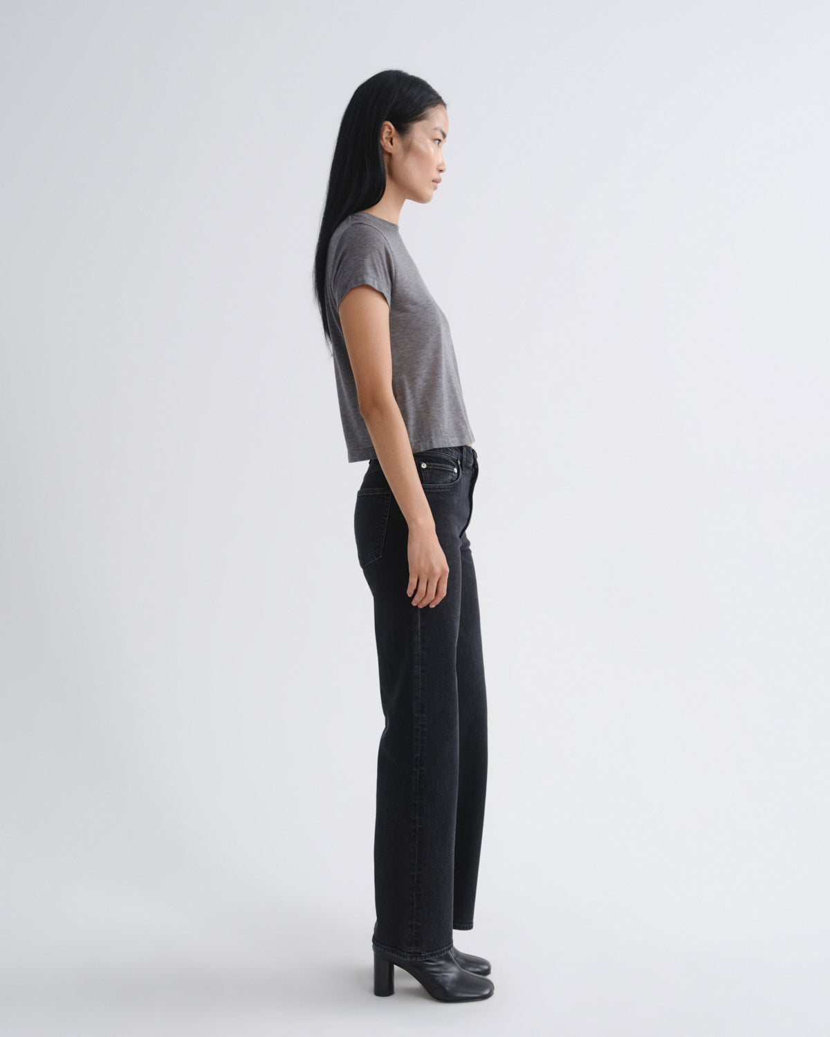 A woman stands in profile wearing a short-sleeve gray knit top, black wide-leg jeans, and black heeled boots against a plain light gray studio background.
