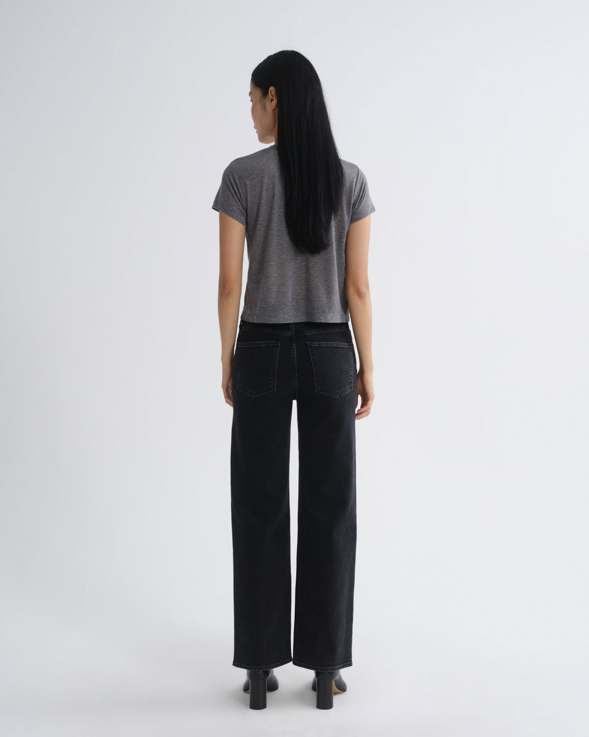 A woman is shown from the back wearing a short-sleeve gray t-shirt and black wide-leg jeans against a plain light gray studio background.