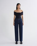 A model stands against a light gray studio background wearing a dark navy off-the-shoulder wrap top, dark blue wide-leg jeans, and black shoes.