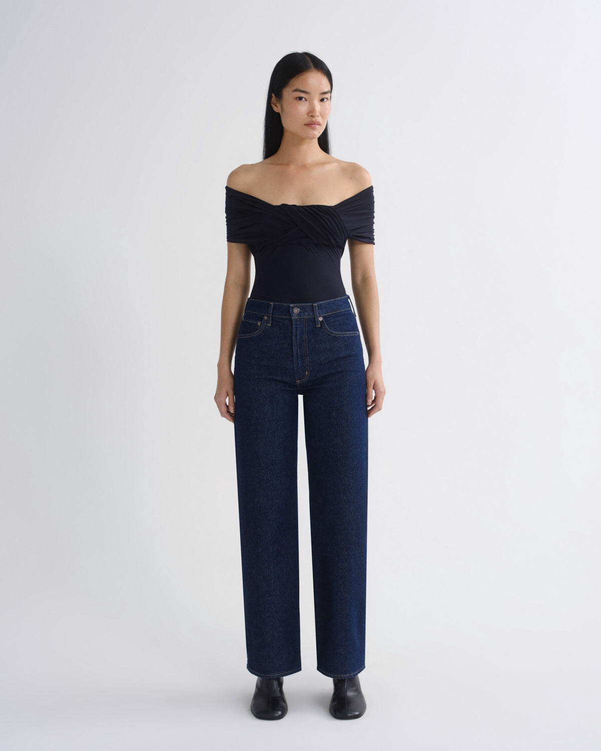 A model stands against a light gray studio background wearing a dark navy off-the-shoulder wrap top, dark blue wide-leg jeans, and black shoes.