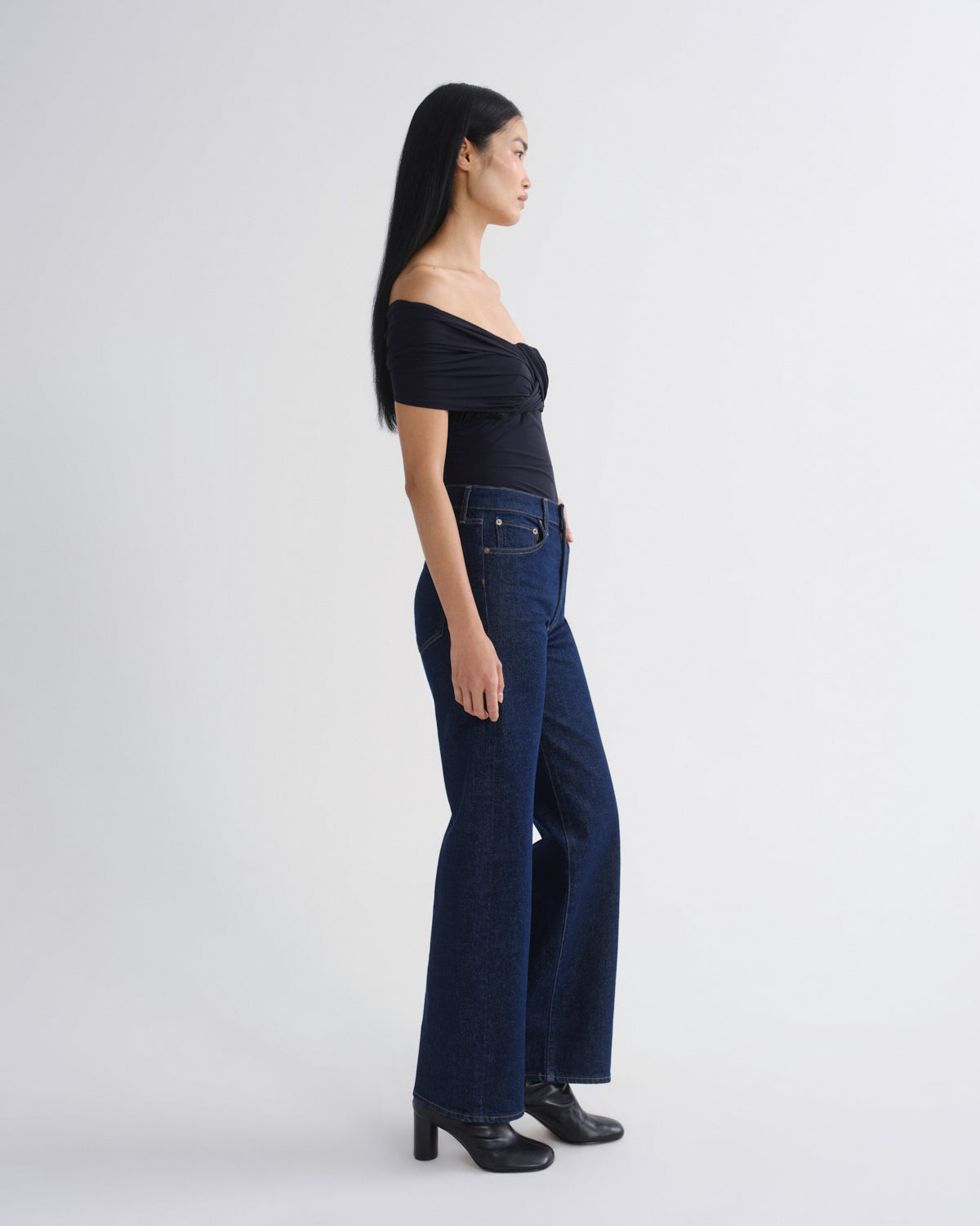 A woman stands in profile wearing a dark off-the-shoulder top, dark blue wide-leg jeans, and black heeled shoes against a plain light gray studio background.